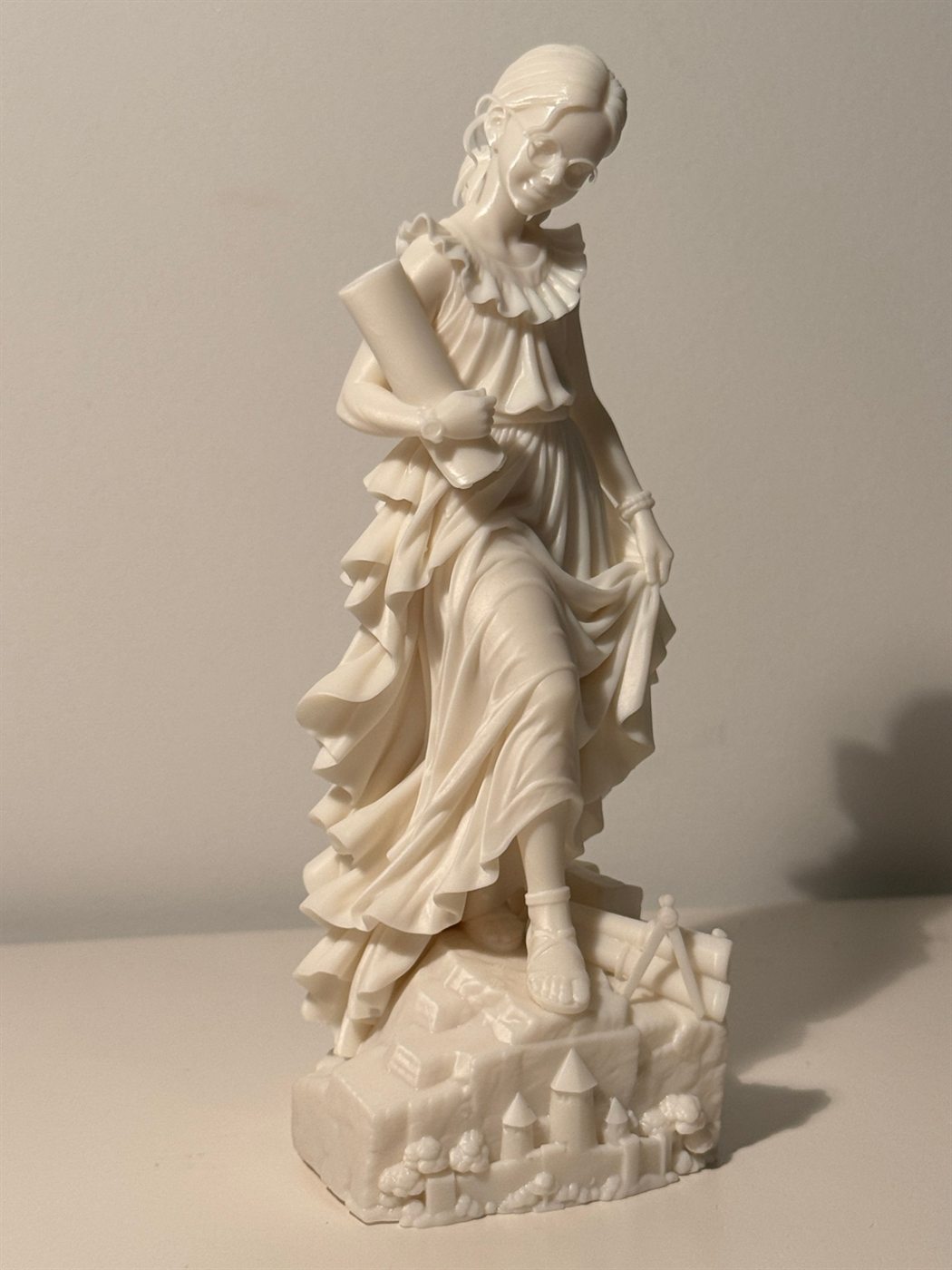 Statuette allégorique en résine blanche : une femme à lunettes en robe drapée, un rouleau de plans sous le bras, le pied posé sur un socle sculpté d'une cité fortifiée